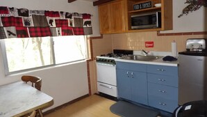 A compact kitchen with a stove, microwave, and sink. There is a dining table and a chair. The kitchen has wooden cabinets and a window with curtains.