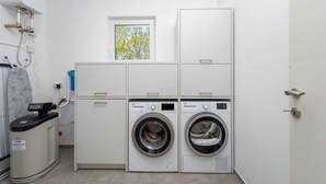 A laundry room with two front-loading washing machines, a drying machine, and a small window with a view of trees. - Villa Larra with heated pool & Jacuzzi (Gostinjac (Krk))