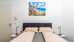 A double bed with a grey mattress and beige pillows, two bedside tables with lamps, a white door, and a painting of a coastal scene on the wall. - Villa Larra with heated pool & Jacuzzi (Gostinjac (Krk))