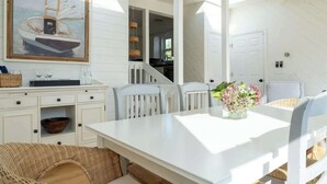 Una cocina moderna con armarios blancos, una mesa de comedor con sillas, una cesta colgante y un cuadro de un barco en la pared.