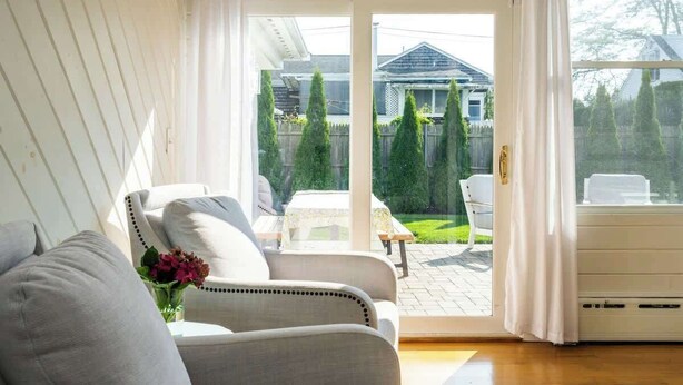Una habitación con puerta corrediza de vidrio que da a un patio con mesa y sillas, un sofá y una pequeña mesa con un florero.