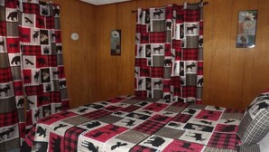 Ein Zimmer mit Holzwandverkleidung, einem Bett mit einer Quiltdecke mit Tiermustern und einem Fenster mit einem rot-schwarz karierten Vorhang.