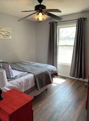 Quarto com cama, ventilador de teto, janela com cortinas e um móvel vermelho.