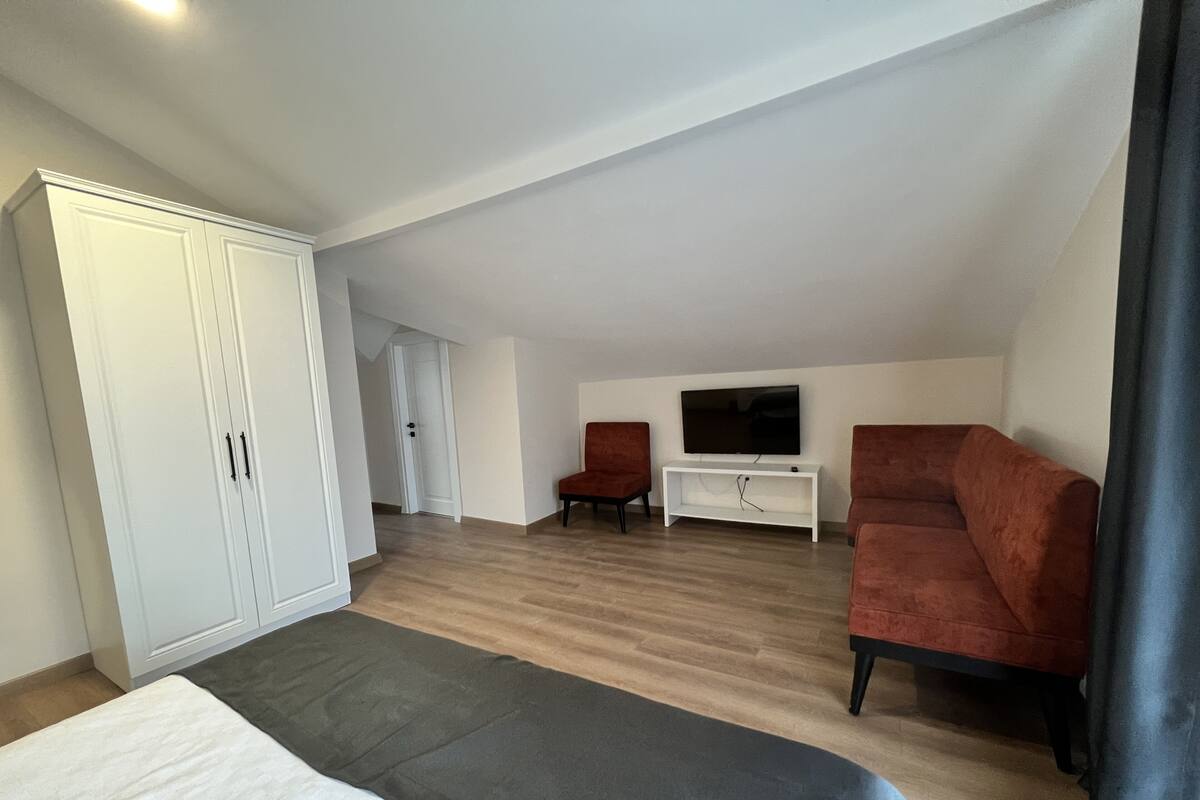 Habitación de hotel con cama, televisor sobre un soporte, dos sillones rojos, armario blanco y puerta que da a otra habitación.
