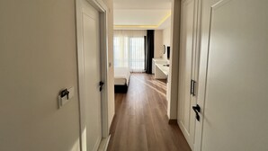 A hotel hallway with wooden flooring, white walls, and doors leading to a bedroom with a bed, a desk, and a TV. - DADA OTEL (Ezine)
