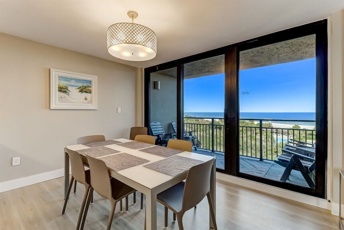 A dining area with a table and chairs, a pendant light, and a large window with a view of the beach and ocean.