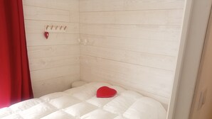 A small, white, wooden-paneled room with a bed, a red heart-shaped pillow, and a wall-mounted hook.