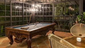 Mesa de bilhar com estrutura de madeira e superfície em feltro azul, envolta em uma proteção de vidro com vista para o verde lá fora.