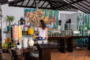 A buffet setup with a woman serving orange juice, tropical decorations, and a mural in the background. - Hotel Campestre Café Café (Montenegro)