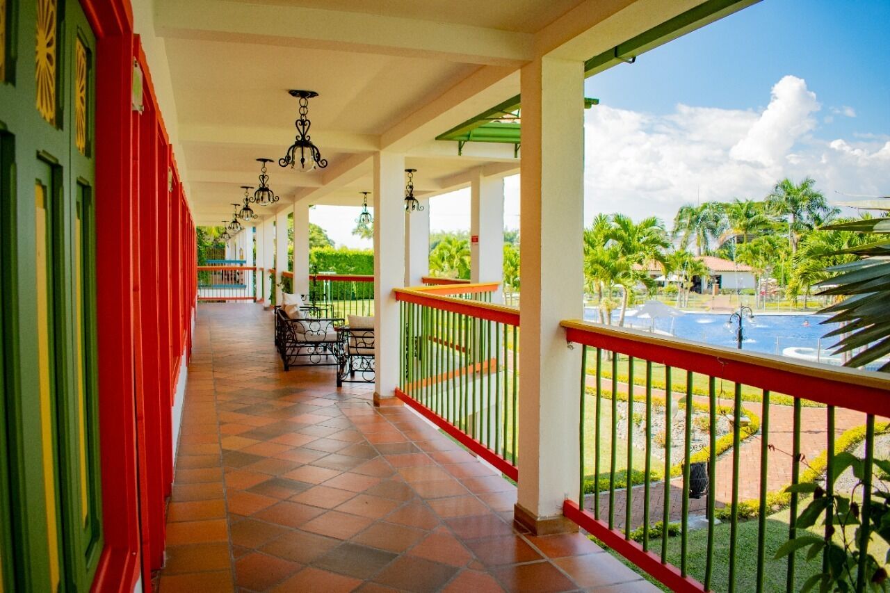 Un espacio al aire libre cubierto con piso de baldosas, barandilla roja y vista a una piscina y palmeras.