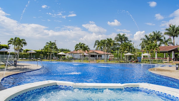 Uma grande piscina ao ar livre com hidromassagem, rodeada por palmeiras e áreas cobertas.