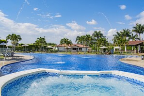 Una gran piscina al aire libre con un jacuzzi, rodeada de palmeras y zonas cubiertas.