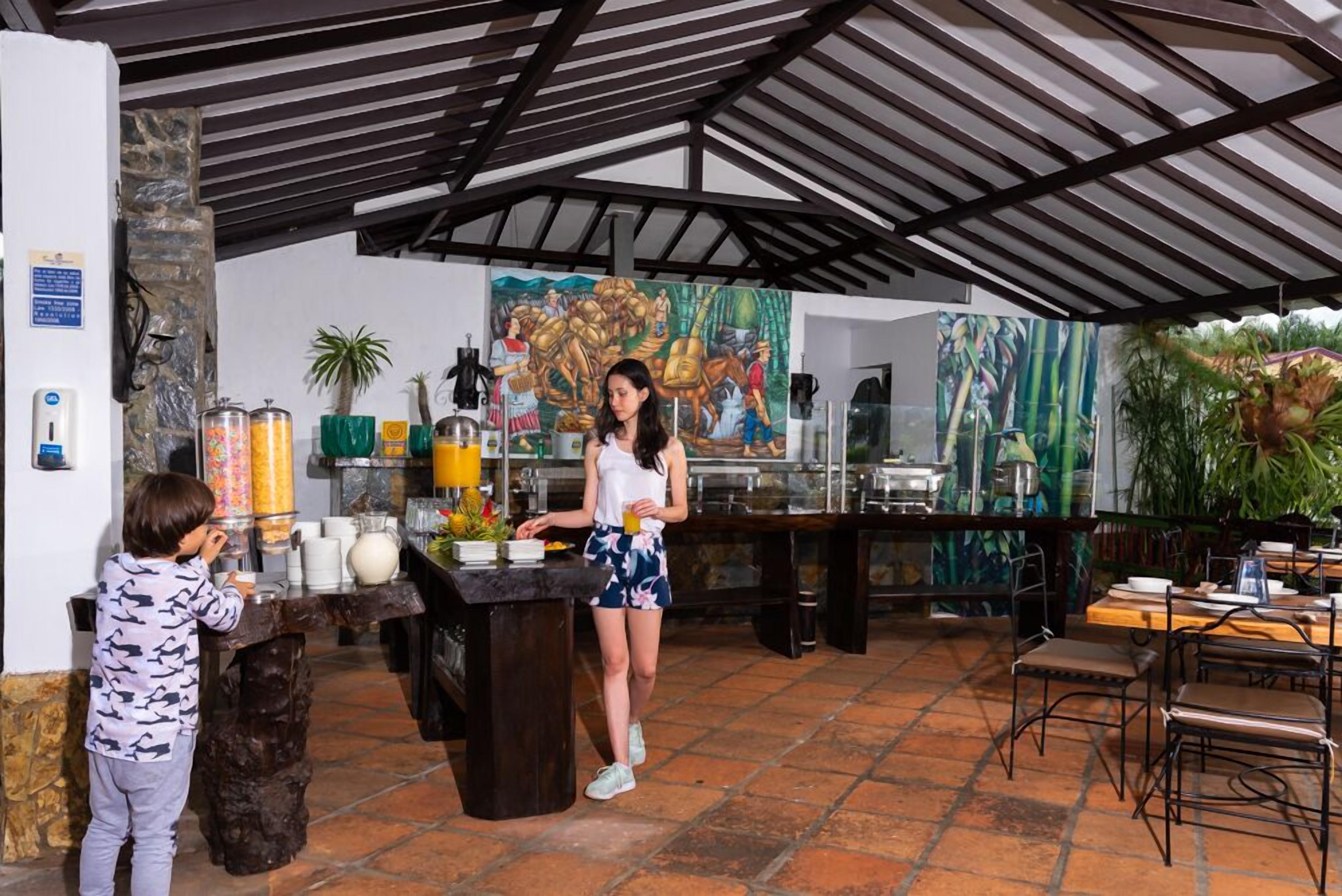 A covered outdoor dining area with a buffet setup, a child interacting with the food station, and a mural in the background.