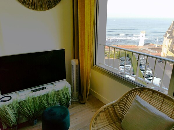 Un balcón con vista al mar, un televisor, un radiador y una silla de mimbre.