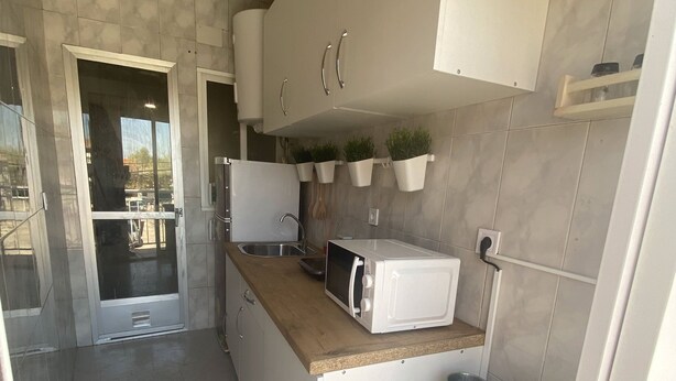 A compact kitchen with white cabinets, a microwave, and a sink. There are hanging plants on the wall and a small storage area with baskets.