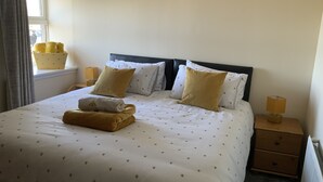 A neatly made bed with a floral-patterned duvet, two pillows, and a folded towel on the bed. There is a nightstand with a lamp and a small wooden chest beside the bed. - Charming Stone Parlour Cottage set in an area of outstanding beauty. (Biggar)