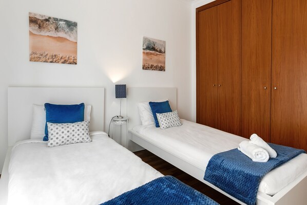A hotel room with two single beds, a wooden wardrobe, and two beach-themed wall art pieces.