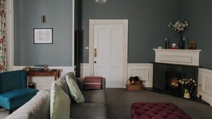 A living room with a grey sofa, teal armchair, and a red ottoman. There is a fireplace with a white mantel, a chandelier, and a framed picture on the wall. - The Pheasant Lodge - Spacious, luxury home with Garden Views & Log fire (Grange-over-Sands)