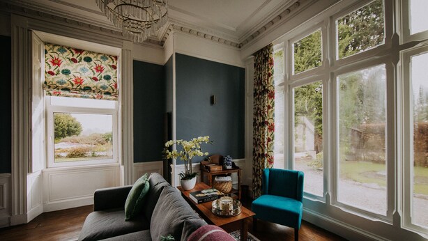 A living room with a sofa, a blue armchair, a wooden coffee table with decorative items, and large windows with floral curtains. - The Pheasant Lodge - Spacious, luxury home with Garden Views & Log fire (Grange-over-Sands)