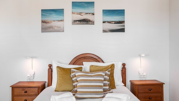 Ein ordentlich eingerichtetes Schlafzimmer mit einem Holz-Kopfteil, einem Bett mit weißen Bettwäsche, zwei Nachttischen mit Lampen und drei gerahmten Bildern an der Wand.