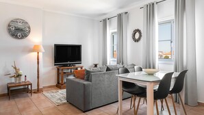 Ein modernes Wohnzimmer mit einem grauen Sofa, einem Fernseher, einem Esstisch mit Stühlen und einem großen Fenster mit Vorhängen.