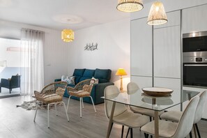 A modern living room with a glass dining table, wicker chairs, a sofa with blue cushions, and a kitchen area with a microwave and oven.