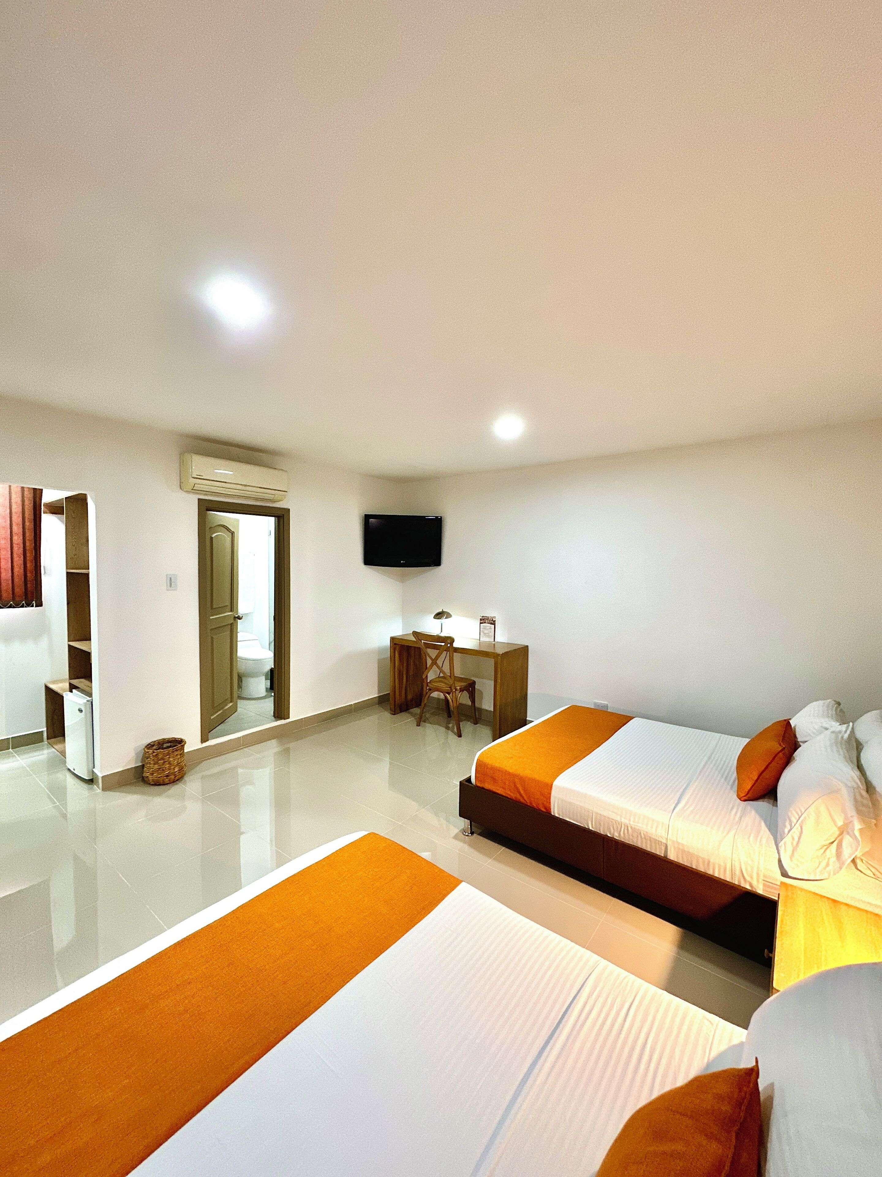 A hotel room with two beds, a desk, a chair, a TV, and a bathroom visible through an open door.
