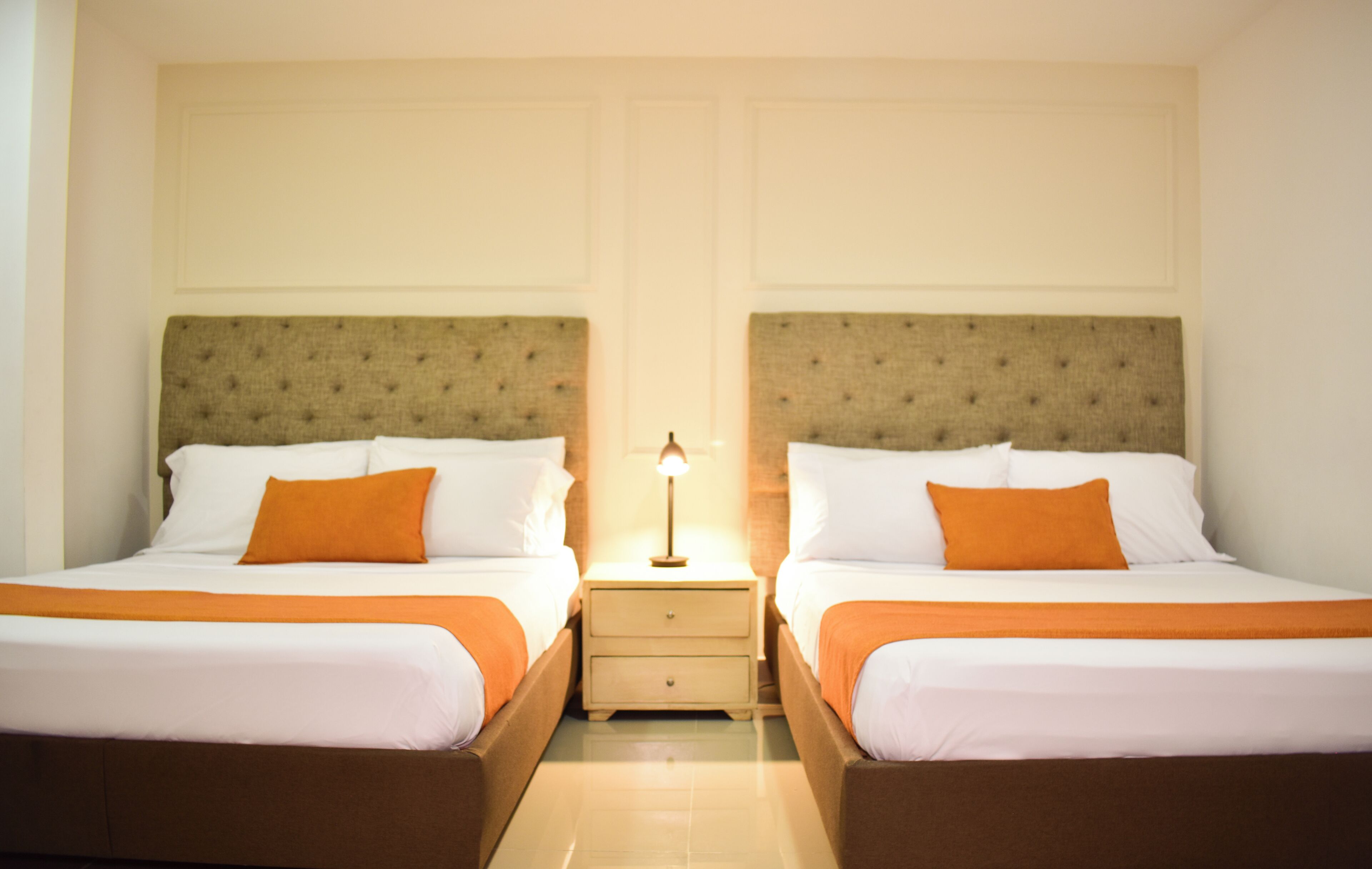 Habitación de hotel con dos camas, cada una con cabecero, ropa de cama blanca y almohadas naranjas. Hay una mesita de noche con una lámpara entre las camas.