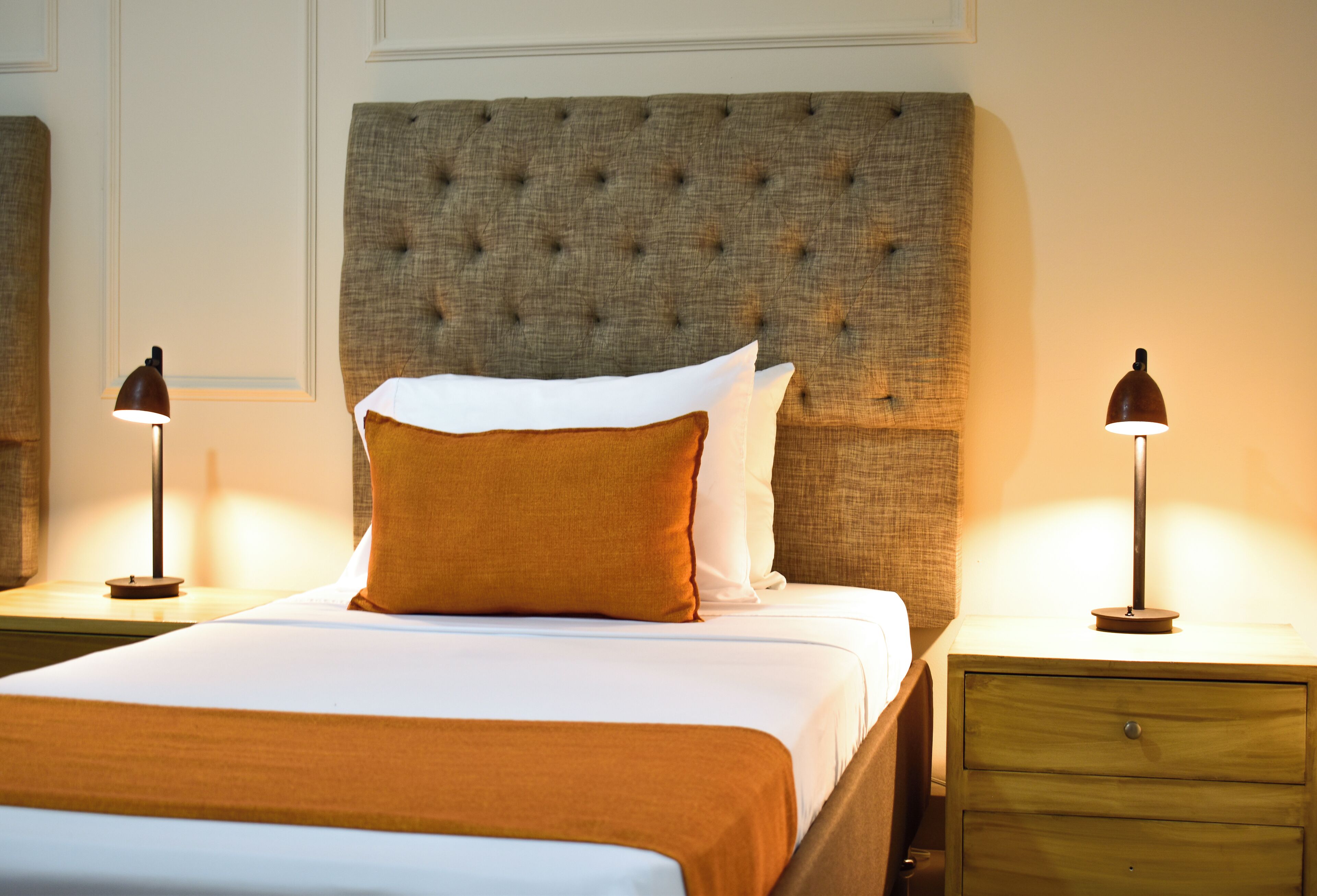 Una cama con ropa de cama bien tendida, un cabecero capitonado, dos mesitas de noche con lámparas y un buró de madera con cajón.