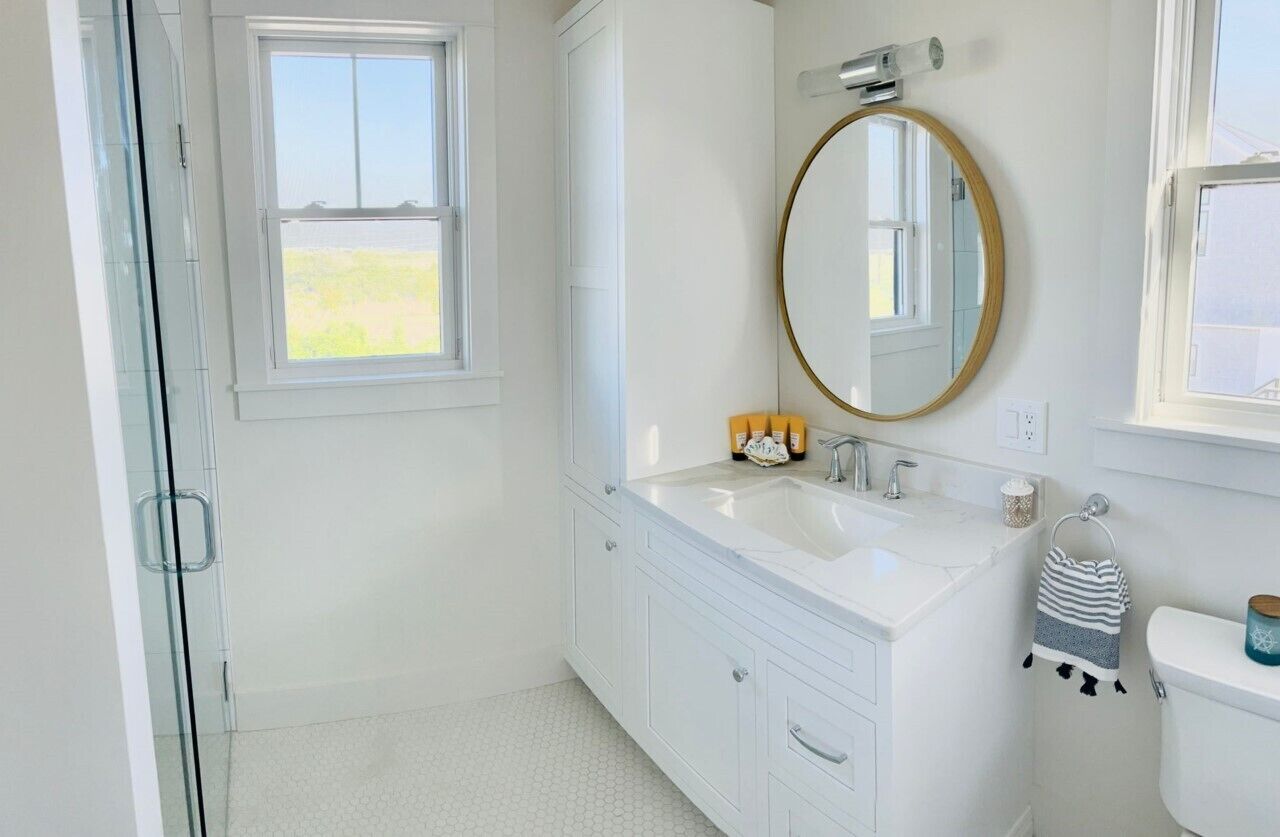 A bathroom with a white vanity, a round mirror, a toilet, and a window.