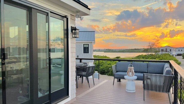 Una terraza moderna con sofá, sillas y mesa, con vistas a un cuerpo de agua al atardecer.