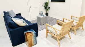 Una sala moderna con un sofá azul oscuro, dos sillas de mimbre, una mesita auxiliar y una planta en maceta.