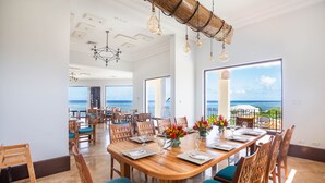 A spacious dining area with a large wooden table set for six, surrounded by wooden chairs with teal cushions. The room features a high ceiling with exposed beams and hanging light fixtures. Large windows offer a view of the ocean and a balcony with outdoor seating. - Pristine Bay Villa 1304  with large pool - 3 bedroom (Roatan)