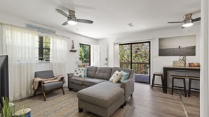 Un salón espacioso con un sofá seccional gris, un sillón pequeño y una mesa de centro de madera. Hay ventiladores de techo, un ventanal con cortinas blancas y vistas al exterior.
