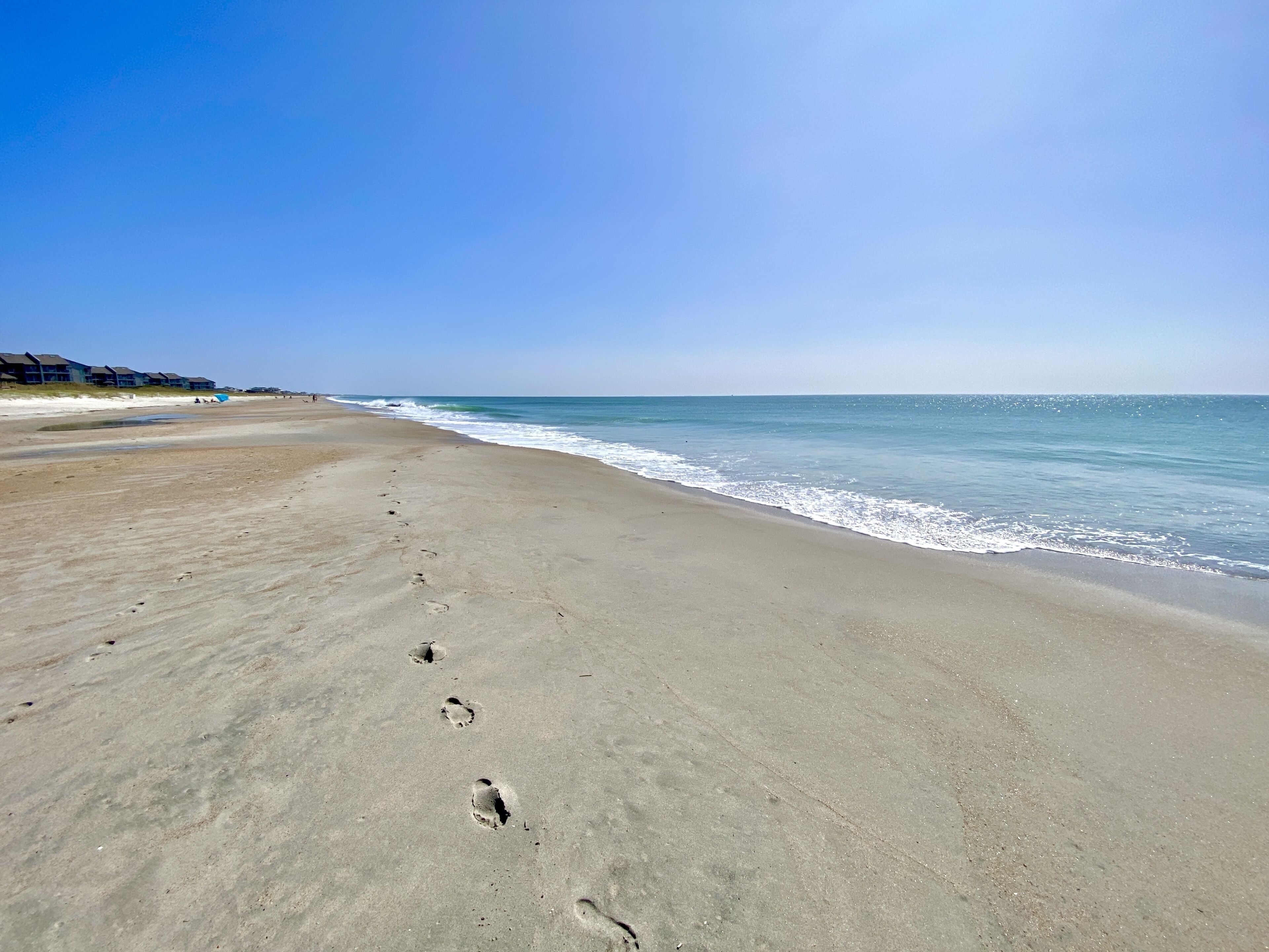 A sandy beach with footprints leading to the water, a clear sky, and buildings in the background.