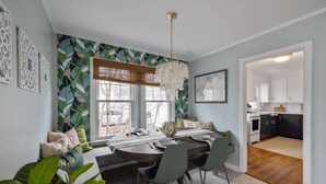 Een eetruimte met een ronde tafel, stoelen en een kroonluchter. Er zijn ramen met jaloezieën, een wand met behang met bladmotieven en uitzicht op de keuken door een open doorgang.