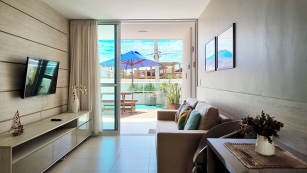 A modern living room with a flat-screen TV, a sofa, a dining table, and a view of an outdoor patio with umbrellas and plants.