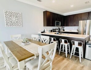 A modern kitchen with dark wood cabinets, a central island with seating, and a dining area with white chairs. - Navarre Beach Front Whole House Large Yard. Kayaks + Paddle Boards Included. (Navarre)