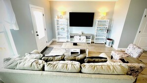A living room with a sofa, coffee table, TV, and shelving units. - Navarre Beach Front Whole House Large Yard. Kayaks + Paddle Boards Included.  (Navarre)