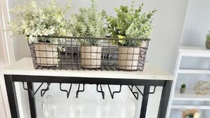A white shelf unit with a black metal frame, holding a variety of items including a watering can, a tray with sticks, and a small plant.
