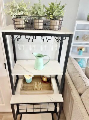 A white shelf unit with a black metal frame, holding a variety of items including a watering can, a tray with sticks, and a small plant.