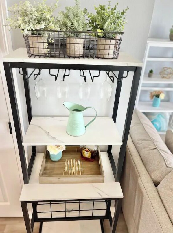 A white shelf unit with a black metal frame, holding a variety of items including a watering can, a tray with sticks, and a small plant.