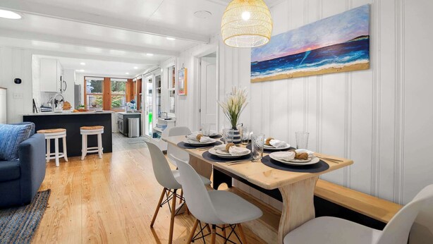 Une cuisine moderne avec des meubles blancs, un coin repas doté d’une table en bois pour quatre personnes, et un grand tableau représentant une plage accroché au mur.