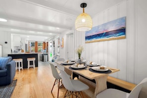 A modern kitchen with white cabinetry, a dining area with a wooden table set for four, and a large beach painting on the wall. - Short Walk to Private Association Beach! (Eastham)