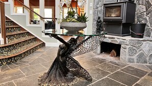 Un interior moderno con una chimenea de piedra, una mesa de vidrio sostenida por una escultura de metal, y una escalera con alfombra estampada.