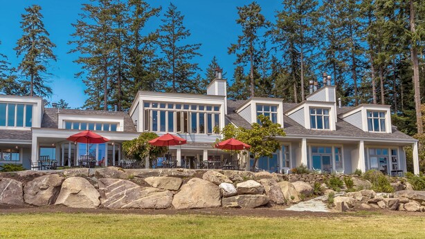 Una casa grande y moderna con varias ventanas, un muro de contención de piedra y sombrillas rojas.