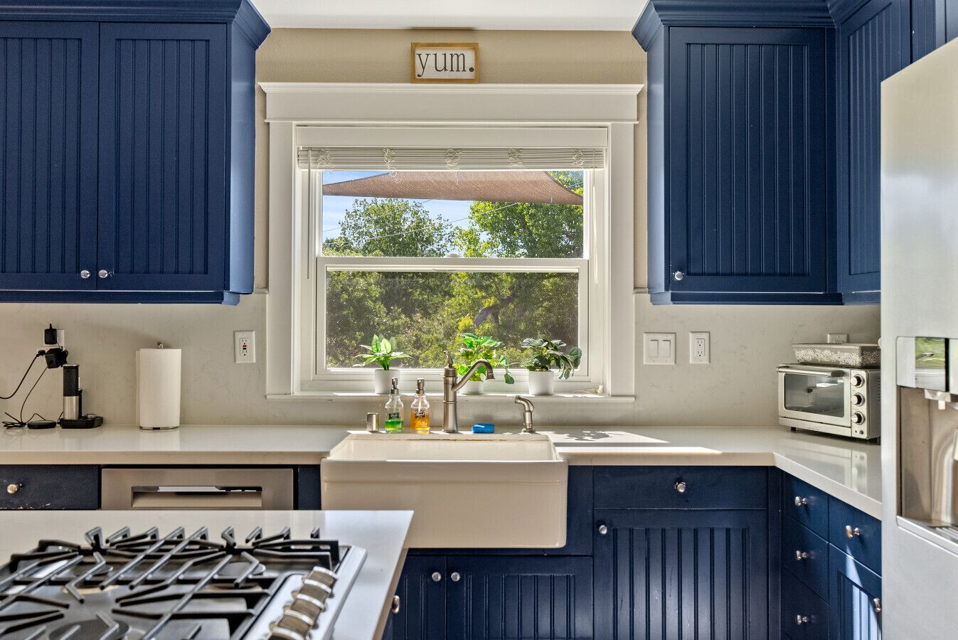 A kitchen with blue cabinets, a window with a view of trees, a sink, a stove, and a toaster.