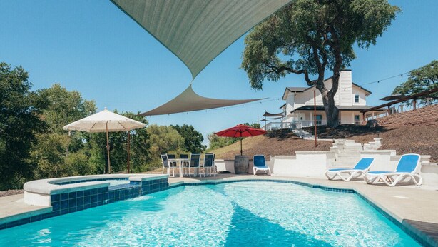 A swimming pool with lounge chairs and umbrellas, surrounded by a landscaped area and a hillside house. - Paso Wine Country Farmhouse | Pool, Spa, Game Room (Paso Robles)