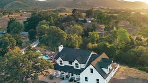 A white two-story house with a dark roof, surrounded by trees and hills. - Paso Wine Country Farmhouse | Pool, Spa, Game Room (Paso Robles)