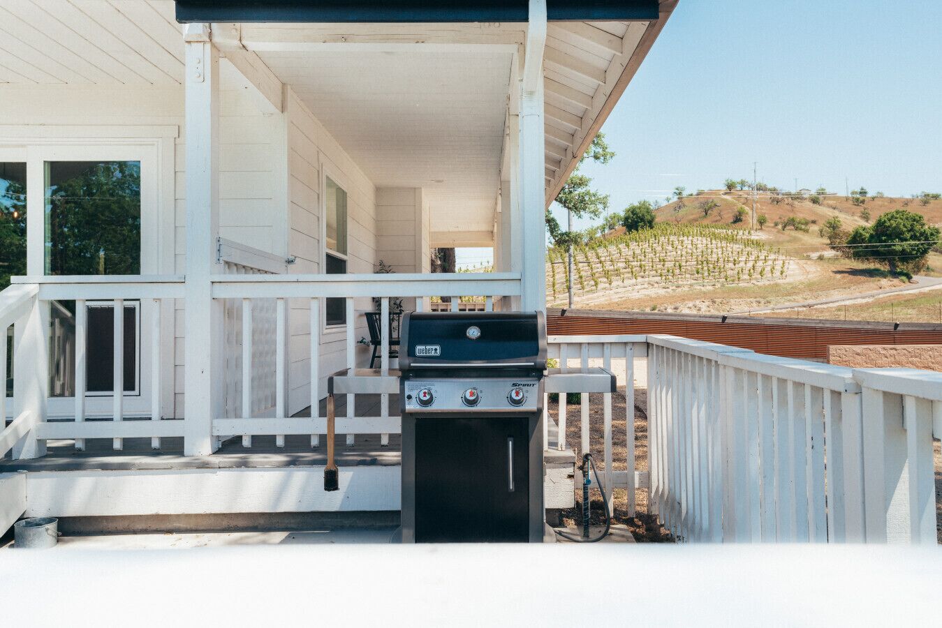 Une véranda couverte avec un barbecue Weber Spirit, une clôture blanche et un vignoble en arrière-plan.